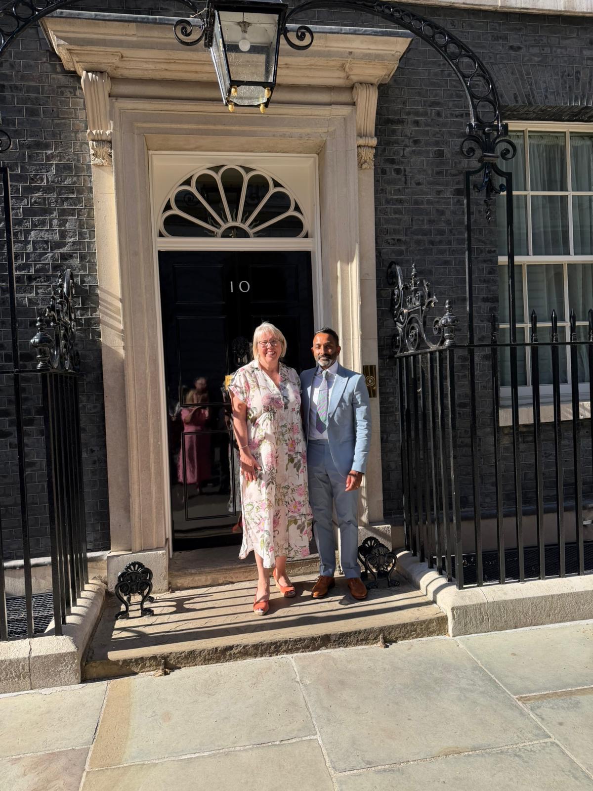 Two people stand on the front steps outside the black door marked "10" at the entrance to a historic brick building. One person is wearing a light-coloured floral dress and the other a pale blue suit with a tie. Ornate black iron railings frame the entrance, and a traditional hanging lantern is positioned above the doorway. Bright sunshine casts strong shadows across the stone steps and pavement in front of the building.