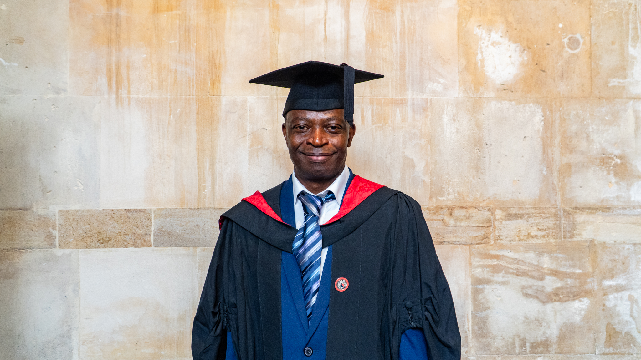 John Adesoye standing in a graduation gown and mortarboard, wearing a blue suit and striped tie, posed against a light stone wall background.