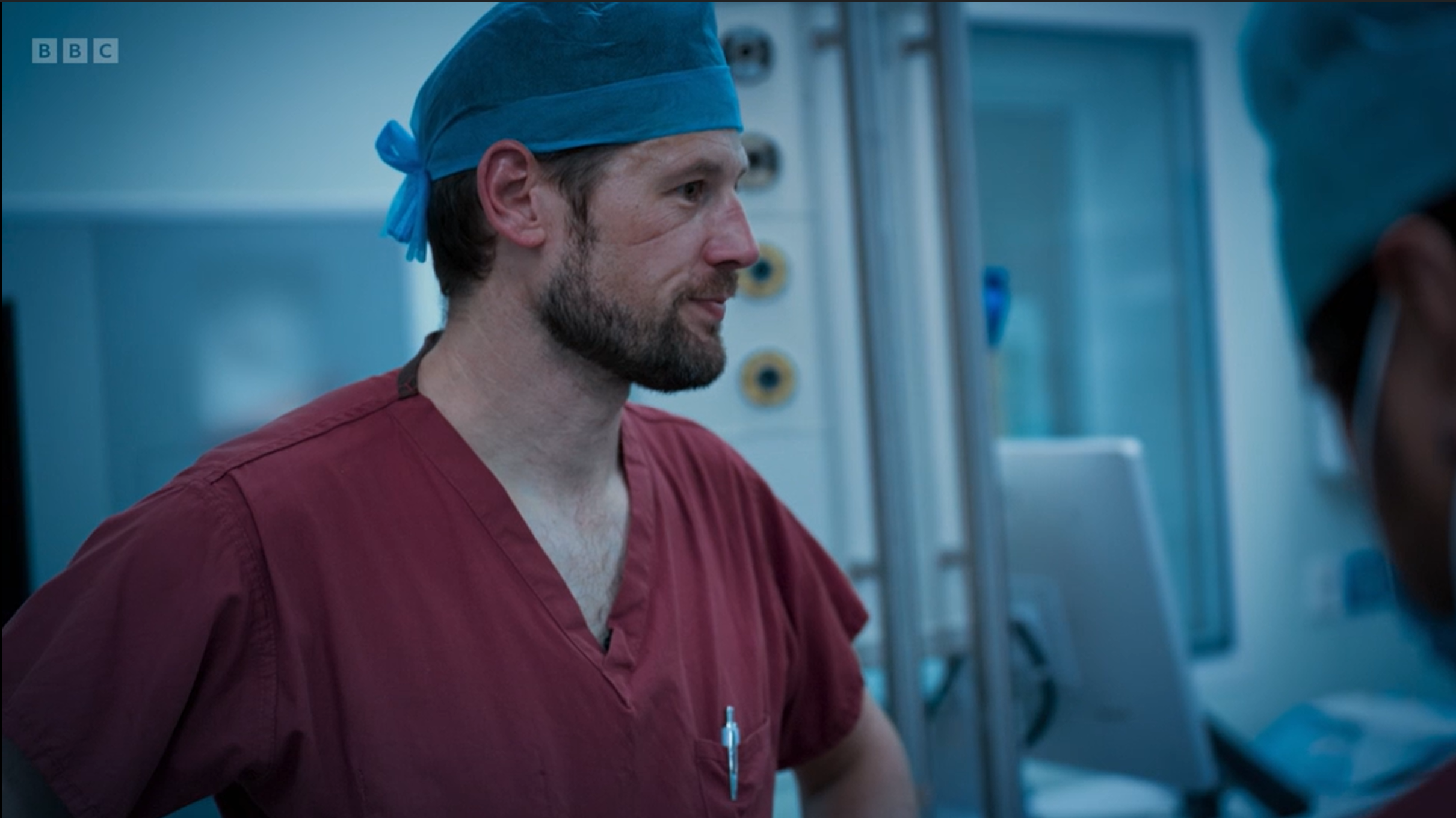 A healthcare professional wearing burgundy scrubs and a blue surgical cap stands in an operating theatre beside medical equipment. The individual is shown in profile, with a blurred colleague in surgical clothing visible in the foreground. The clinical setting is lit with cool blue tones, and monitoring equipment can be seen in the background.