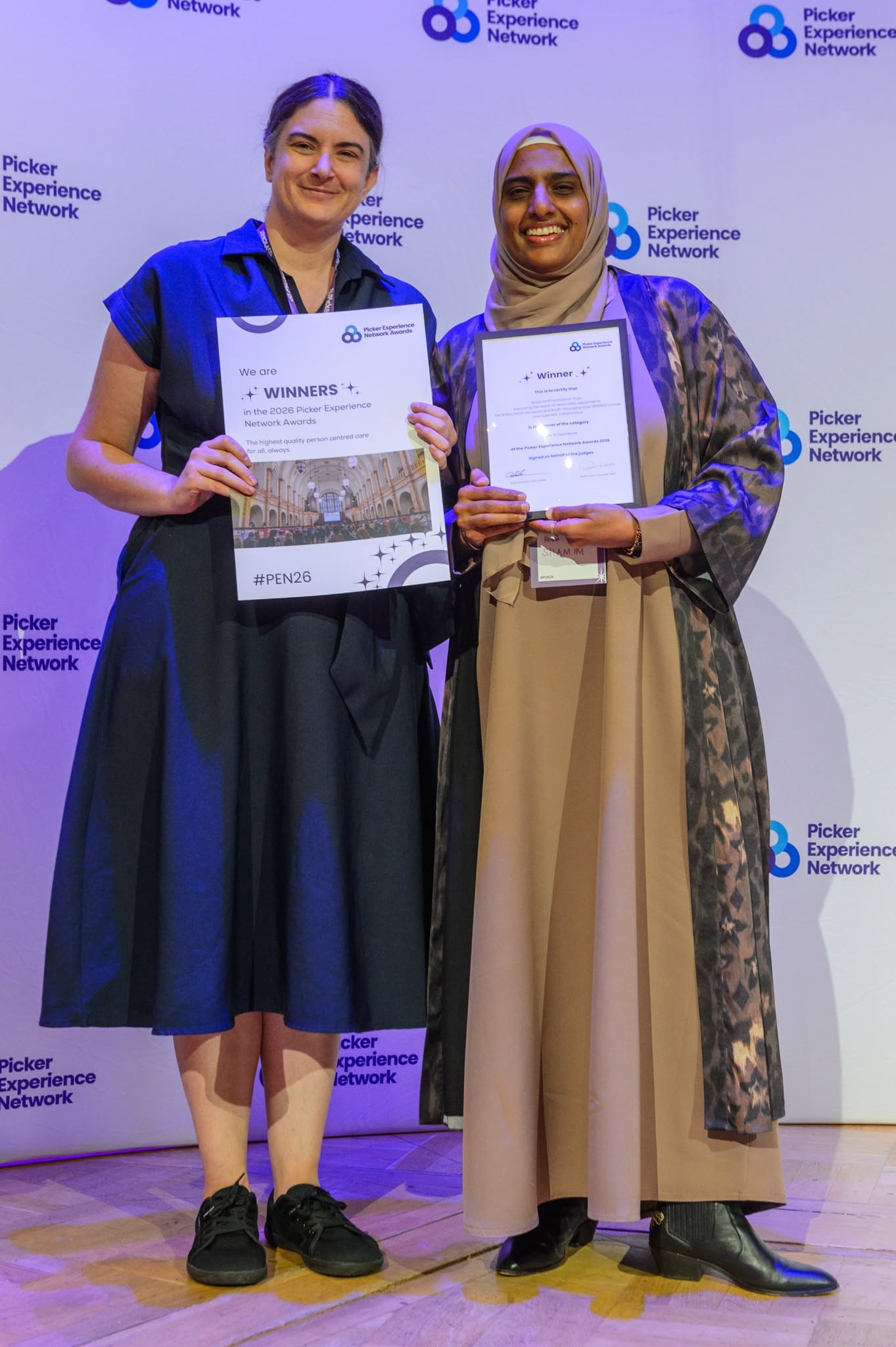 A woman in a navy dress and one in a hijab, smiling and holding certificates that read "winners"