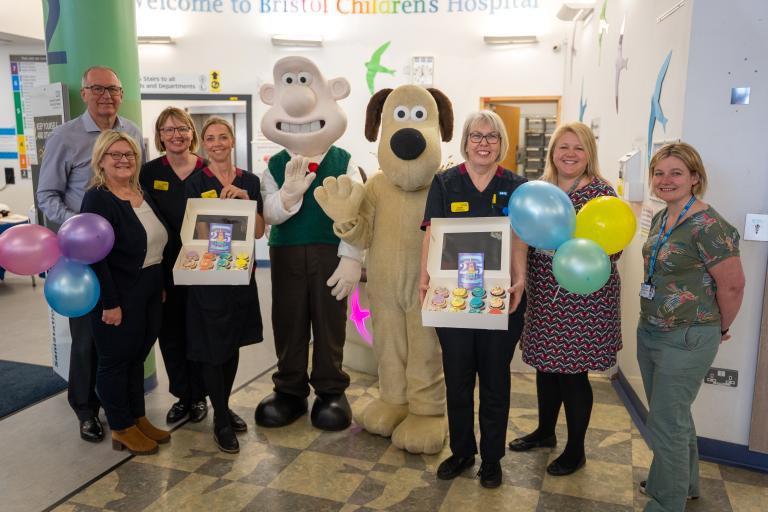 Divisional leaders pose with Wallace and Gromit characters at Bristol Royal Hospital for Children’s 25th birthday celebration, holding cupcakes and balloons.