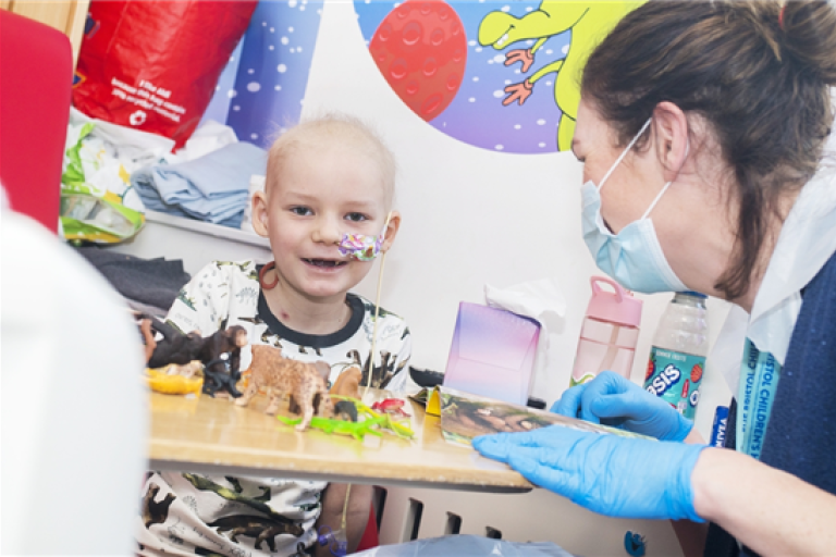 Young patient smiles at a play table with animal toys, interacting with a masked healthcare worker