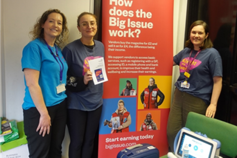 Jade King, Peer Support Lead at the Hepatitis C Trust, Sally Tilden, UHBW Hepatology Clinical Nurse Specialist and Annie Archer, UHBW Hepatology Registrar, posing next to a colourful Big Issue display banner, with a small medical device on chair in foreground