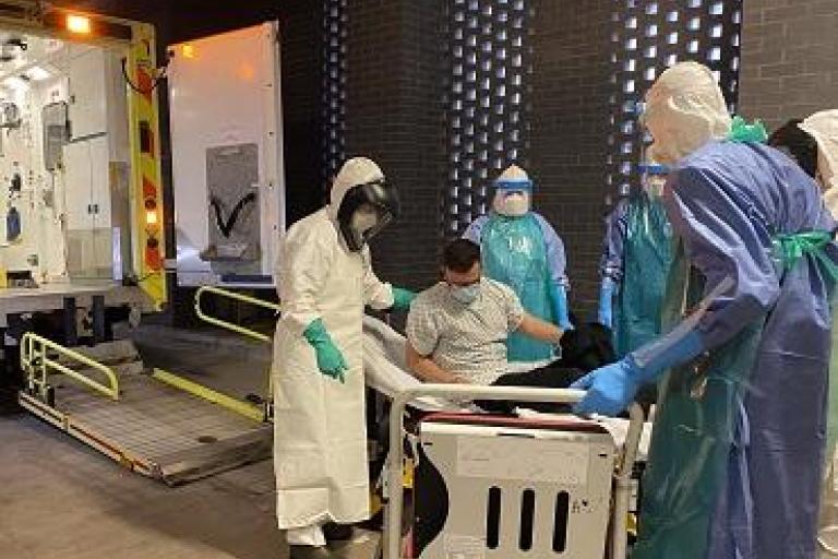 Simulation of a high-consequence infectious disease patient transfer with staff in protective suits assisting a seated patient into an ambulance