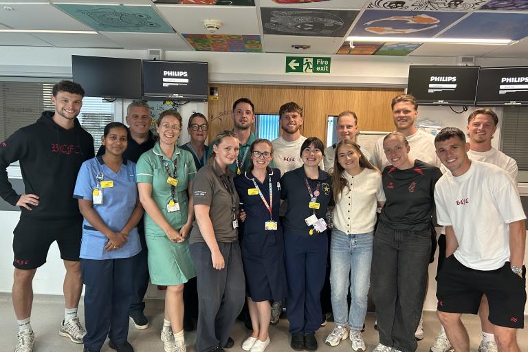 A group of healthcare staff and visitors pose together for a photo inside a hospital ward beneath a ceiling decorated with colourful artwork.