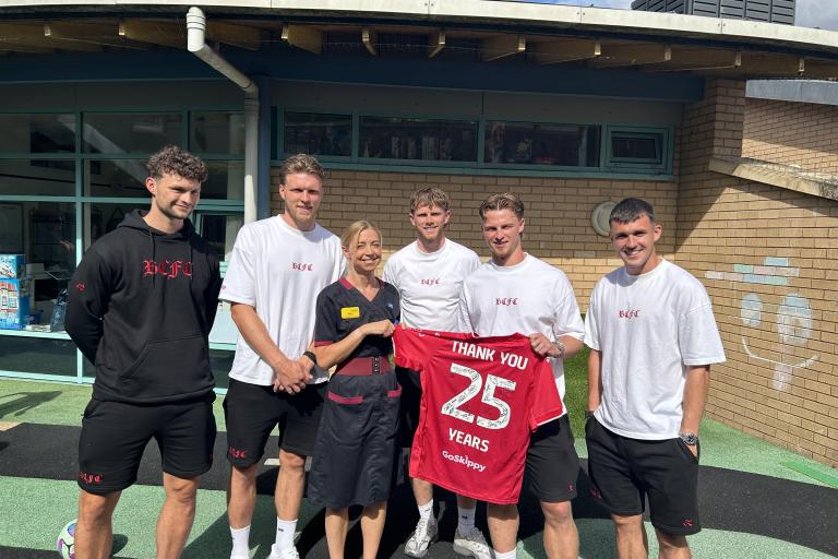 Five visitors and a healthcare staff member stand together in a hospital garden, holding a signed red football shirt that reads “Thank You 25 Years."