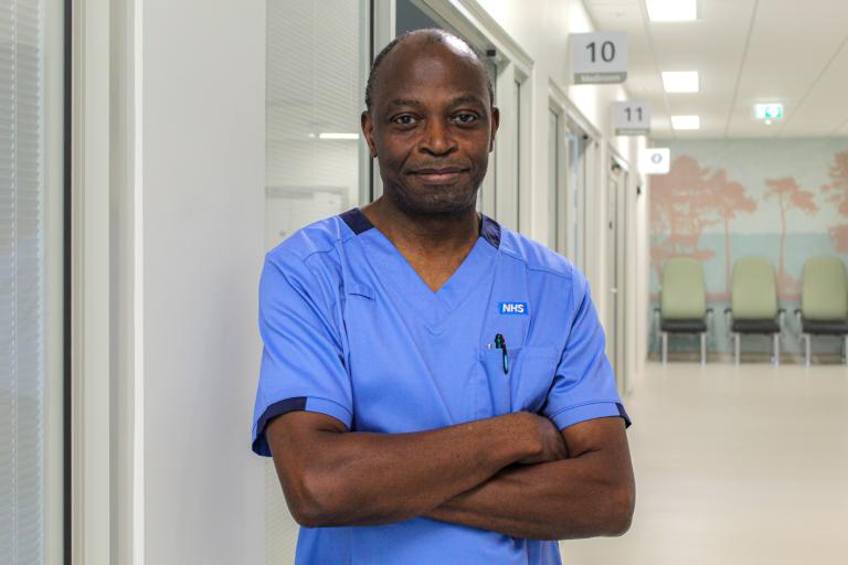 John Adesoye wearing blue NHS nurse uniform posing arms crossed in the Princess Royal Surgical Centre Medirooms