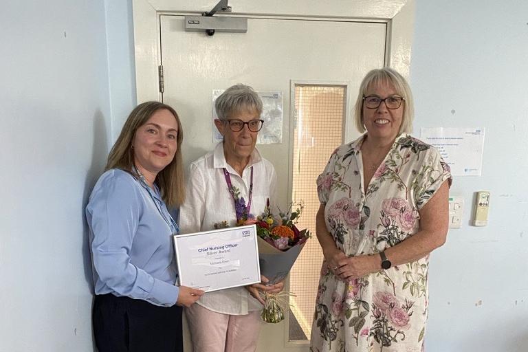 Michaela Dixon stands next to Cora Pressley from NHS England and a colleague holding her CNO award and flowers