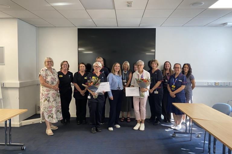 A group of people stand together in a room in front of black screen with two people holding certificates for their chief nurse officer awards