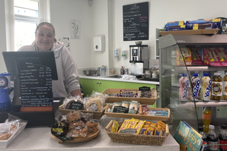A person stood behind a counter, food and drink items are in a fridge to the right of the picture.