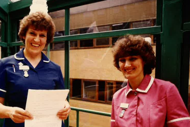 Two nurses, one in blue management uniform and another in pink staff nurse uniform stand together in front of Weston General Hospital on opening day