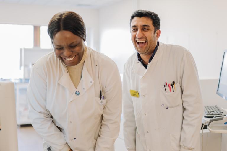 Two healthcare laboratory staff members in white coats working together beside laboratory equipment in a bright clinical workspace.