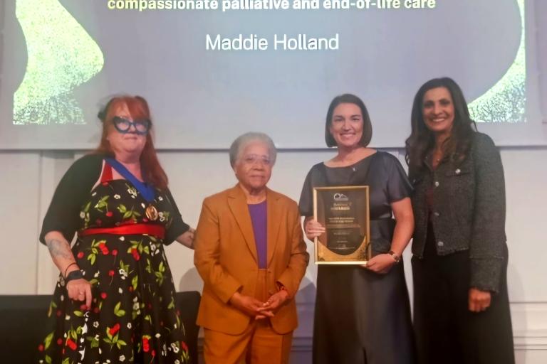 Four people stand on a stage during an awards ceremony. One person holds a framed award plaque while standing beside three other presenters. A large screen behind them displays the text: “Trevor Clay Award – For outstanding achievement in advancing compassionate palliative and end-of-life care.” The recipient’s name, “Maddie Holland,” is shown on the screen above the group. The individuals pose for a photograph in front of the presentation screen.