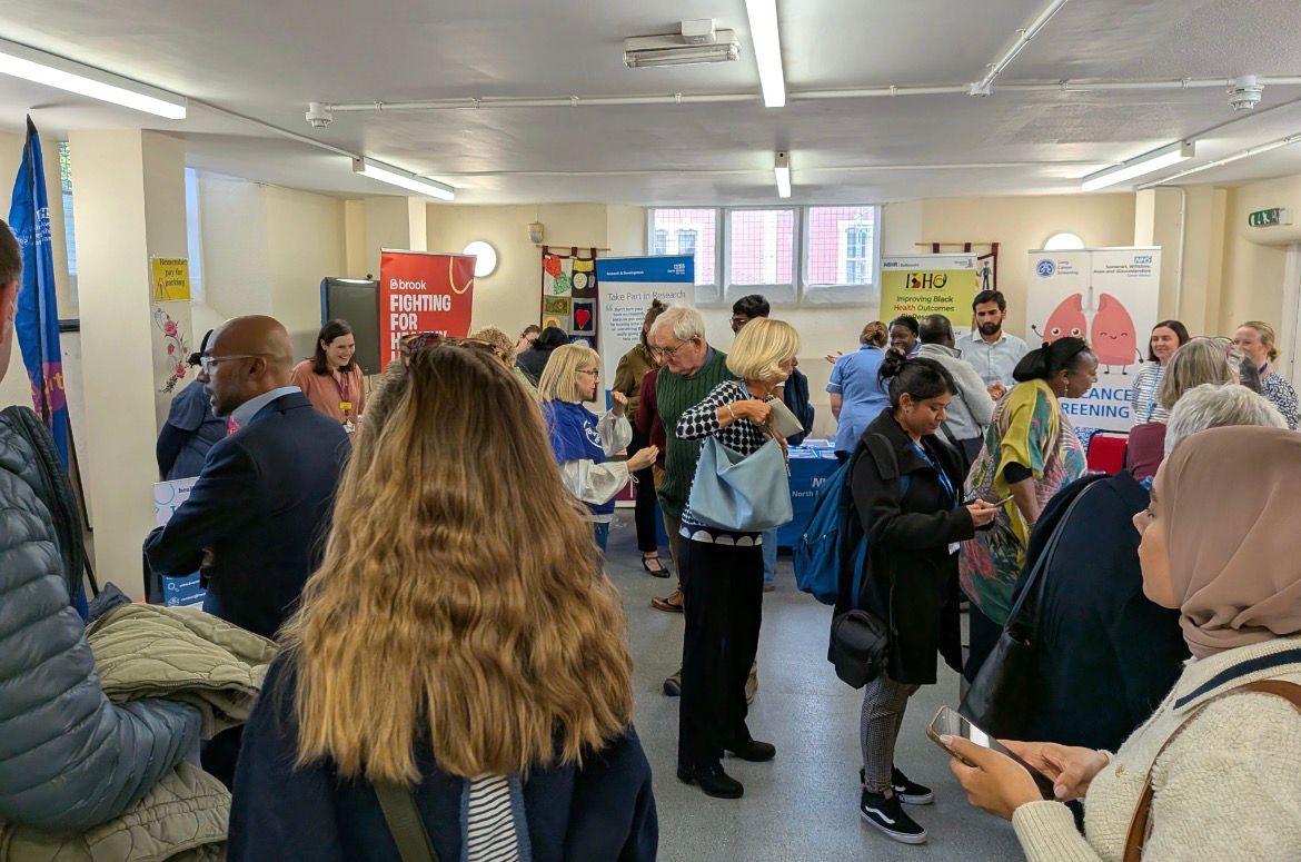 Lots of people are gathered in a room looking at information stands, and talking to each other.