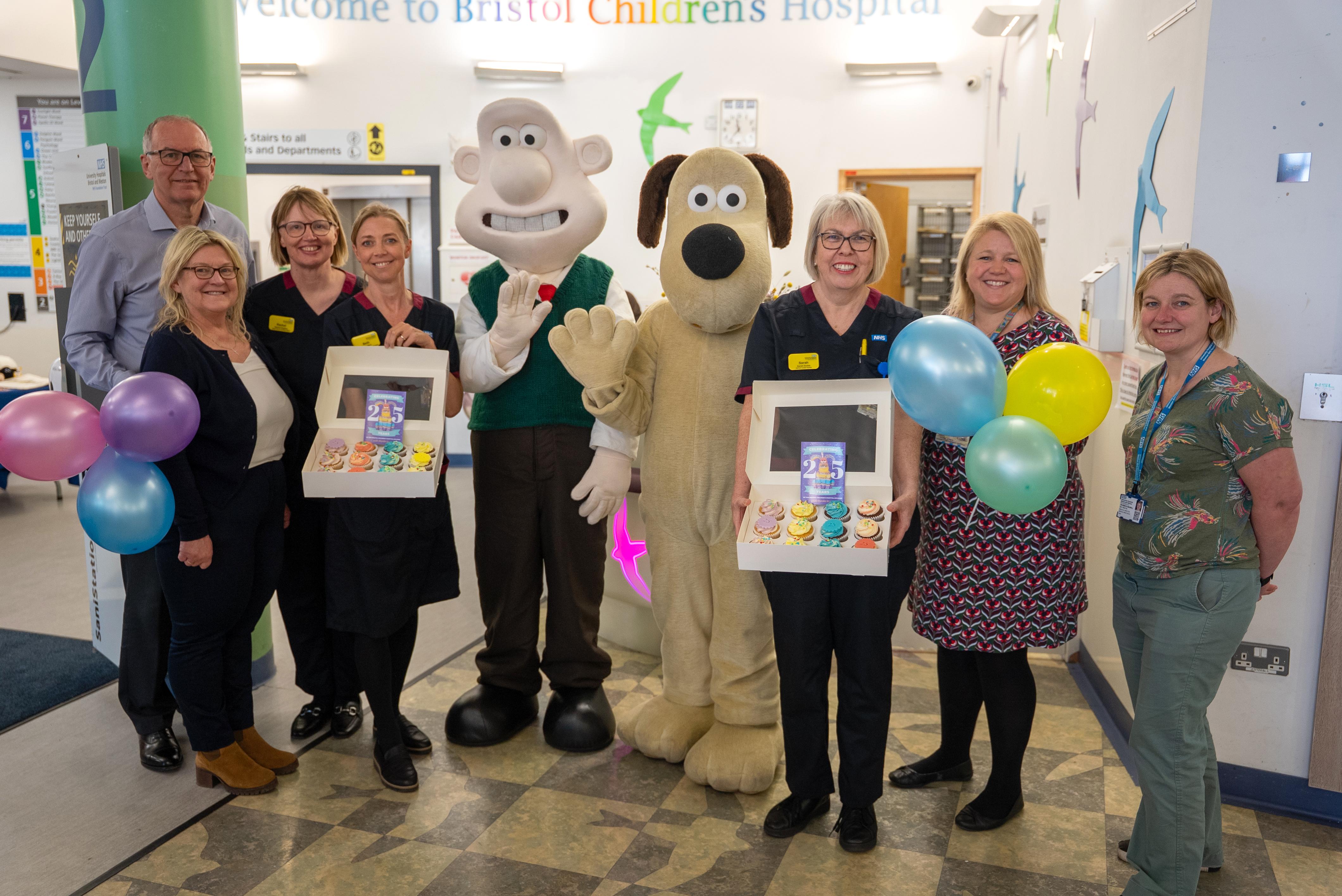 Divisional leaders pose with Wallace and Gromit characters at Bristol Royal Hospital for Children’s 25th birthday celebration, holding cupcakes and balloons.