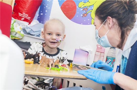 Young patient smiles at a play table with animal toys, interacting with a masked healthcare worker