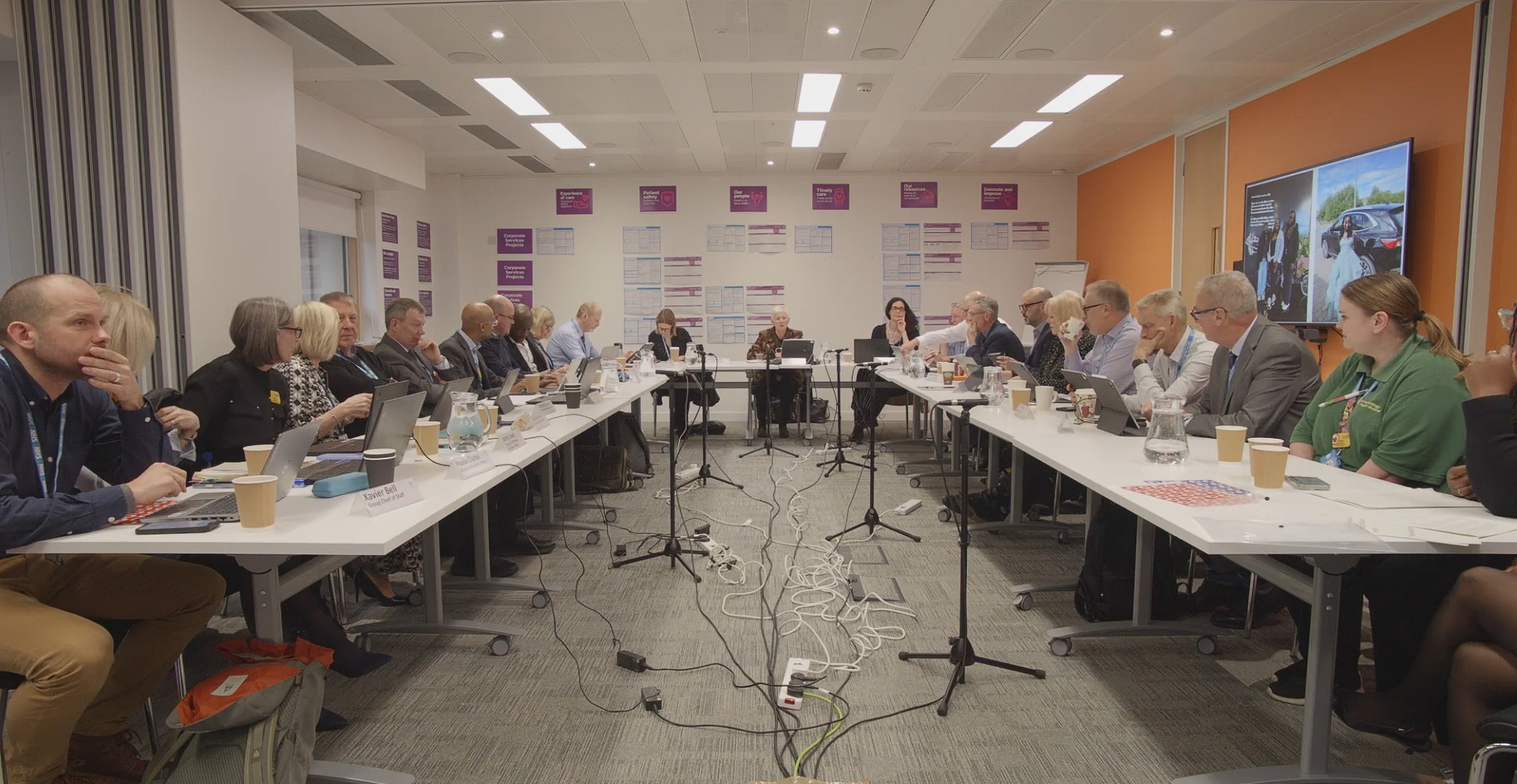 A wide shot of a room with several people sat around tables facing each other. There are several microphones on stands in the middle of the room.