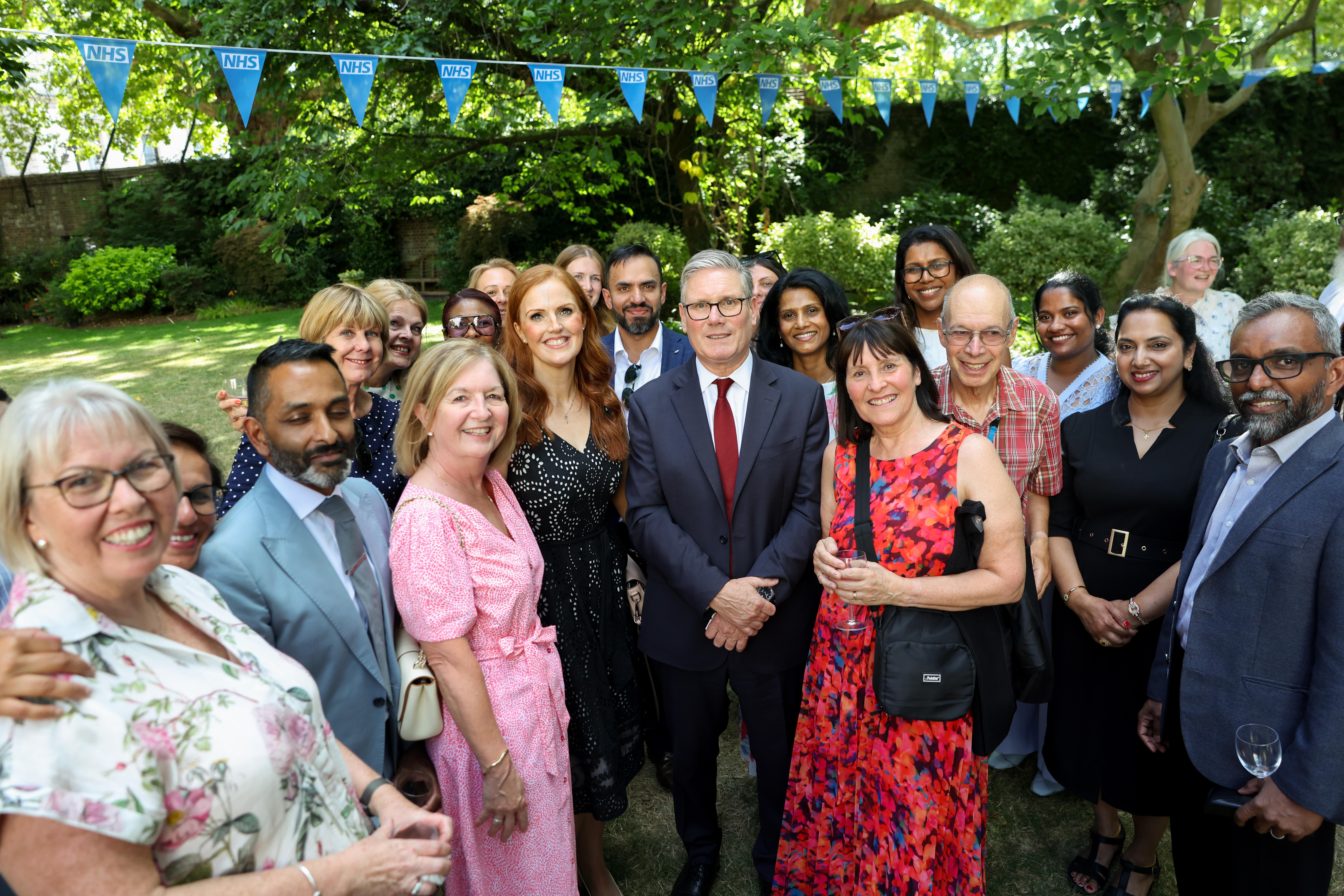 A large group of people stand together for a photograph in a leafy garden during an outdoor NHS event. Blue NHS bunting is strung between trees overhead. The group is arranged closely together on a lawn, with several people wearing formal or semi-formal clothing, including suits, dresses and smart shirts. Shrubs, lawn and mature trees form the background, and some attendees are holding drinks. The image captures a group gathering in a relaxed garden setting beneath NHS-branded decorations.