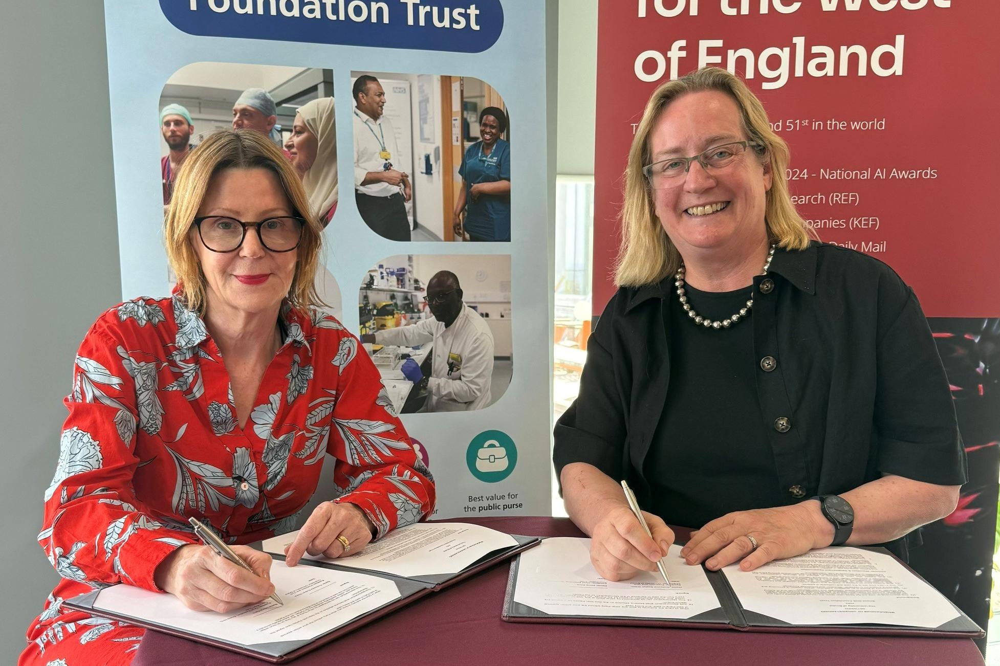 Maria Kane and Prof Evelyn Welch signing the formal memorandum of understanding in front of Bristol NHS Foundation Trust and University of Bristol stand up banners