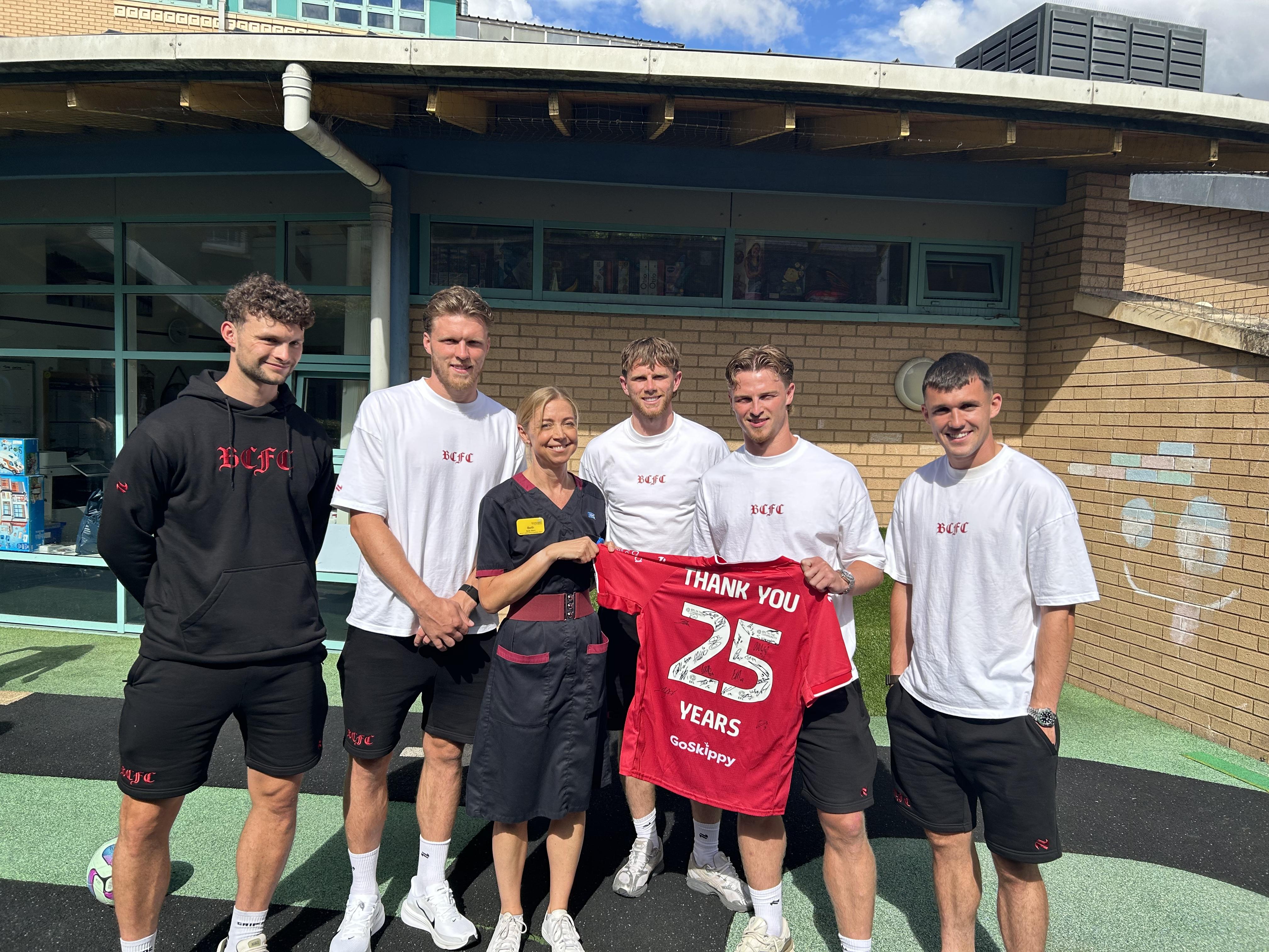 Five visitors and a healthcare staff member stand together in a hospital garden, holding a signed red football shirt that reads “Thank You 25 Years."