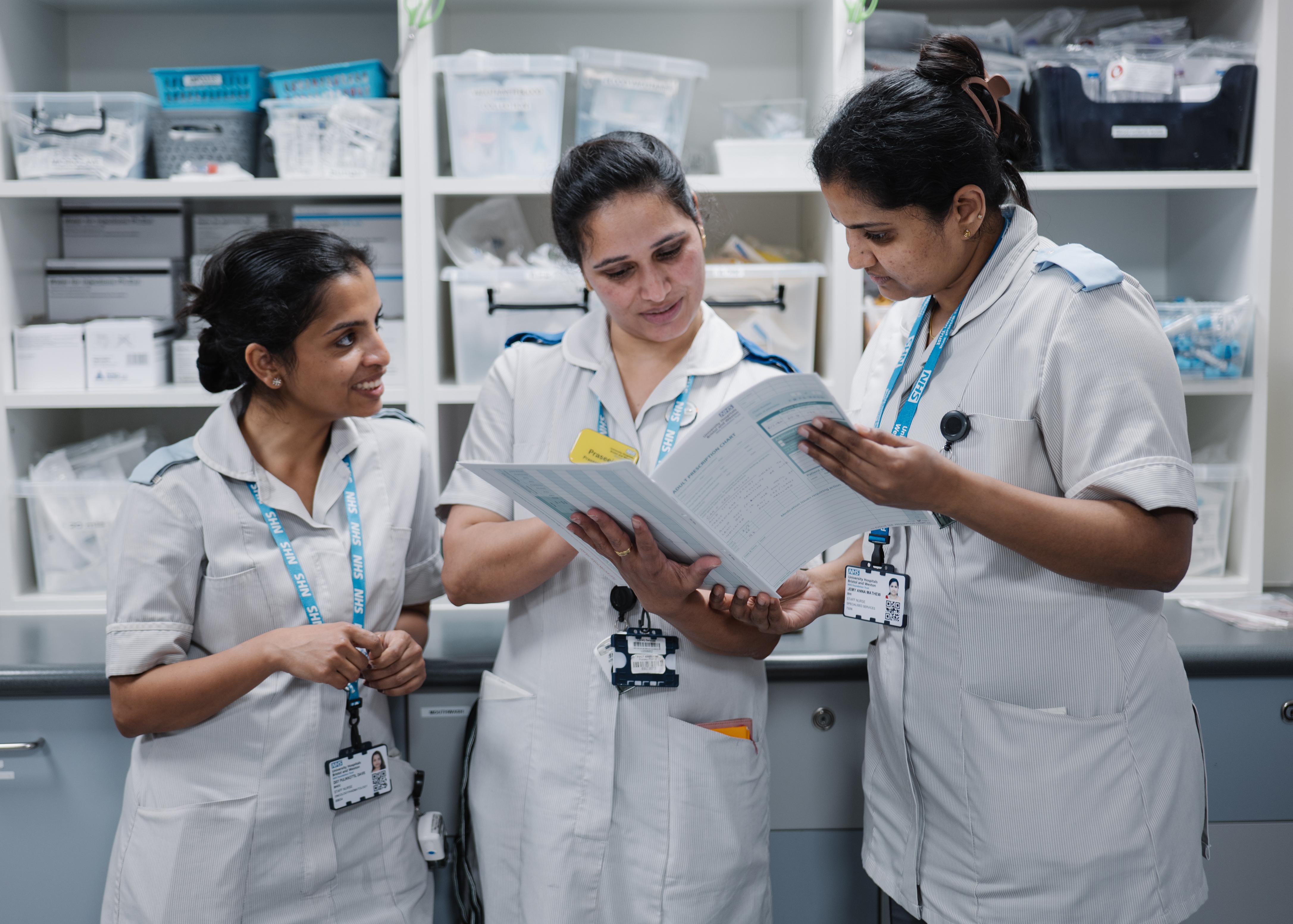 Three nurses from diverse backgrounds, all in uniform, stood together and looking at a file of patient notes. 