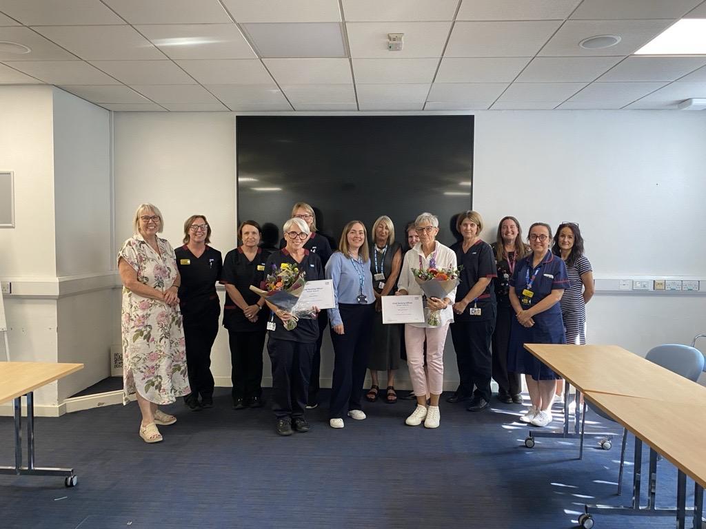 A group of people stand together in a room in front of black screen with two people holding certificates for their chief nurse officer awards