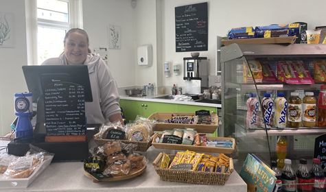 A person stood behind a counter, food and drink items are in a fridge to the right of the picture.