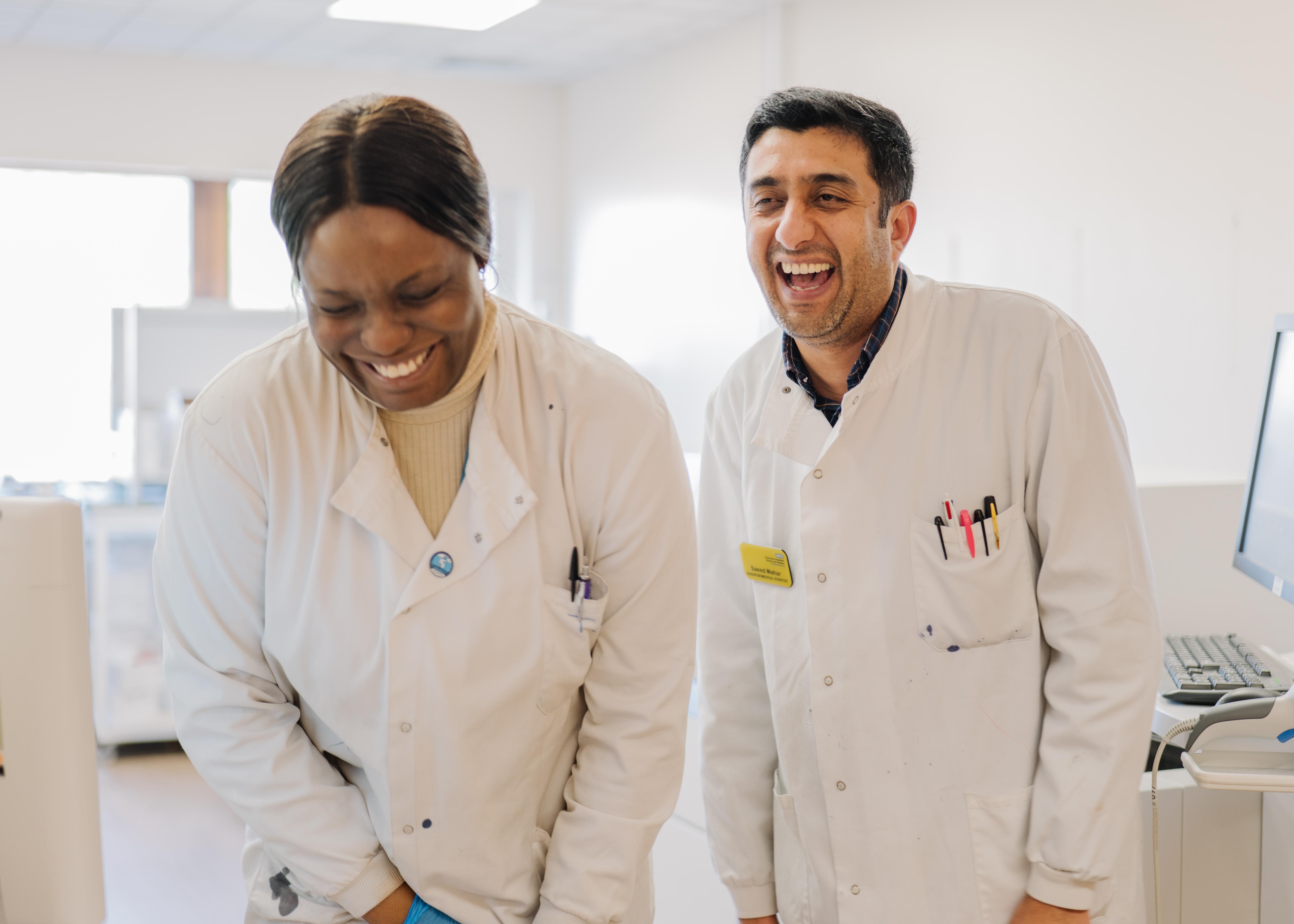 Two healthcare laboratory staff members in white coats working together beside laboratory equipment in a bright clinical workspace.