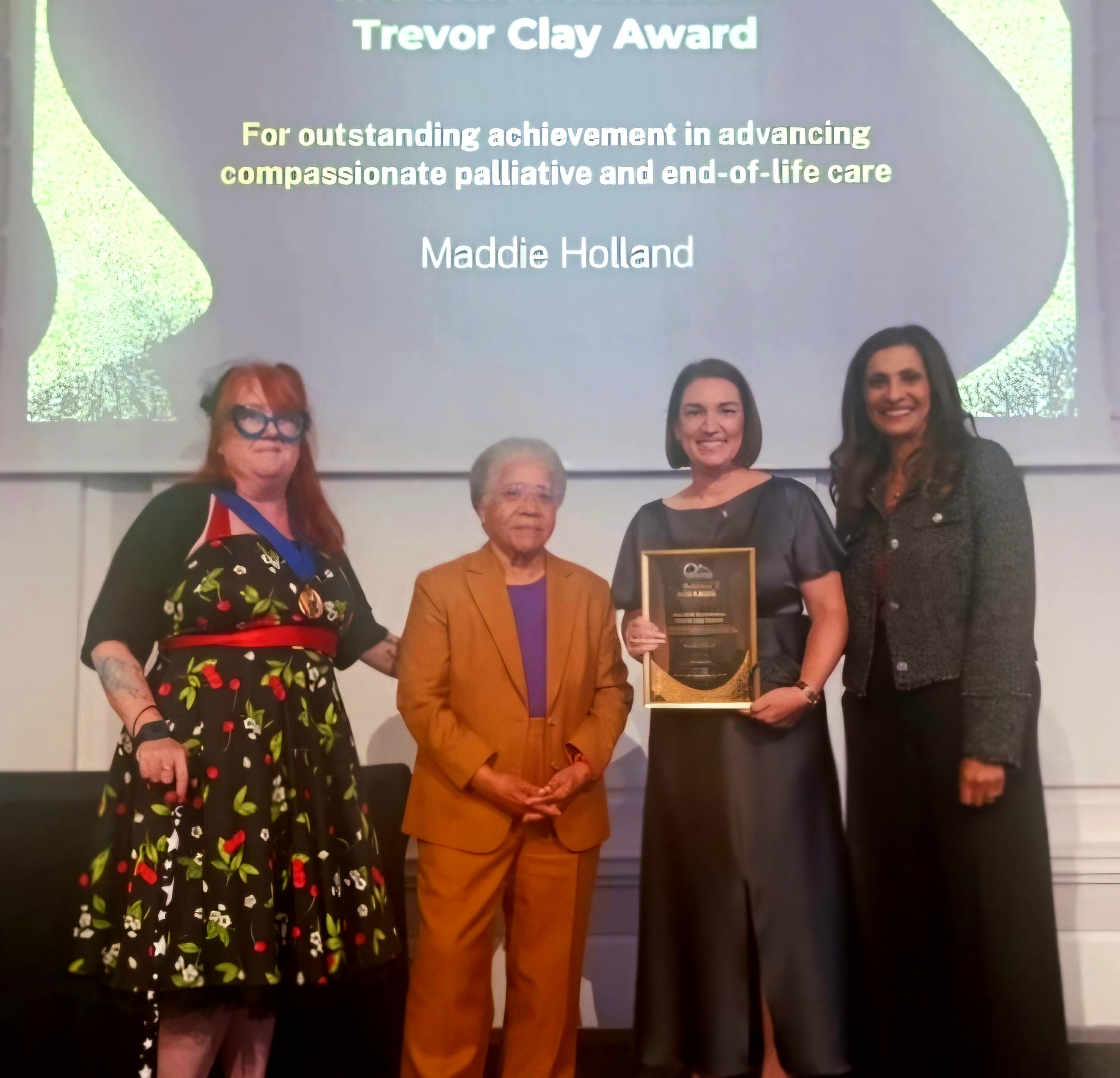 Four people stand on a stage during an awards ceremony. One person holds a framed award plaque while standing beside three other presenters. A large screen behind them displays the text: “Trevor Clay Award – For outstanding achievement in advancing compassionate palliative and end-of-life care.” The recipient’s name, “Maddie Holland,” is shown on the screen above the group. The individuals pose for a photograph in front of the presentation screen.