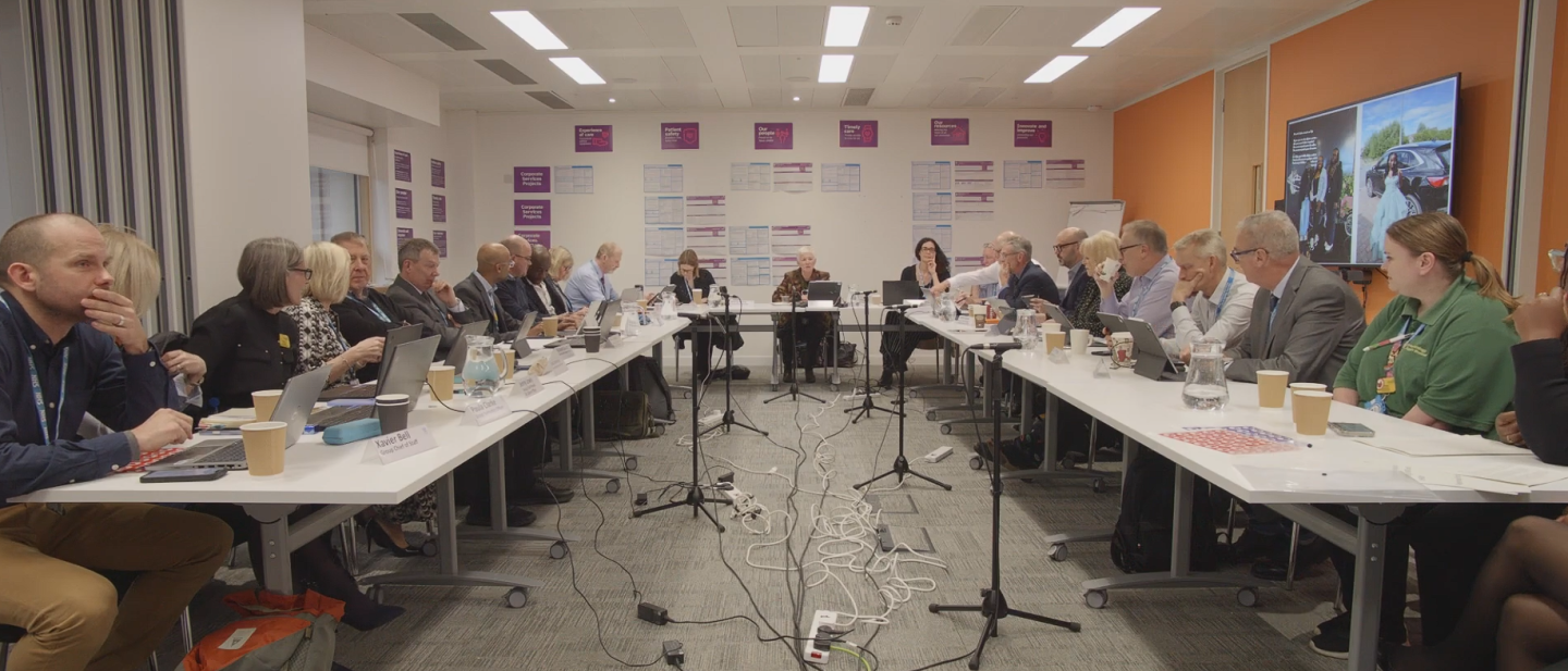 A wide shot of a room with several people sat around tables facing each other. There are several microphones on stands in the middle of the room.
